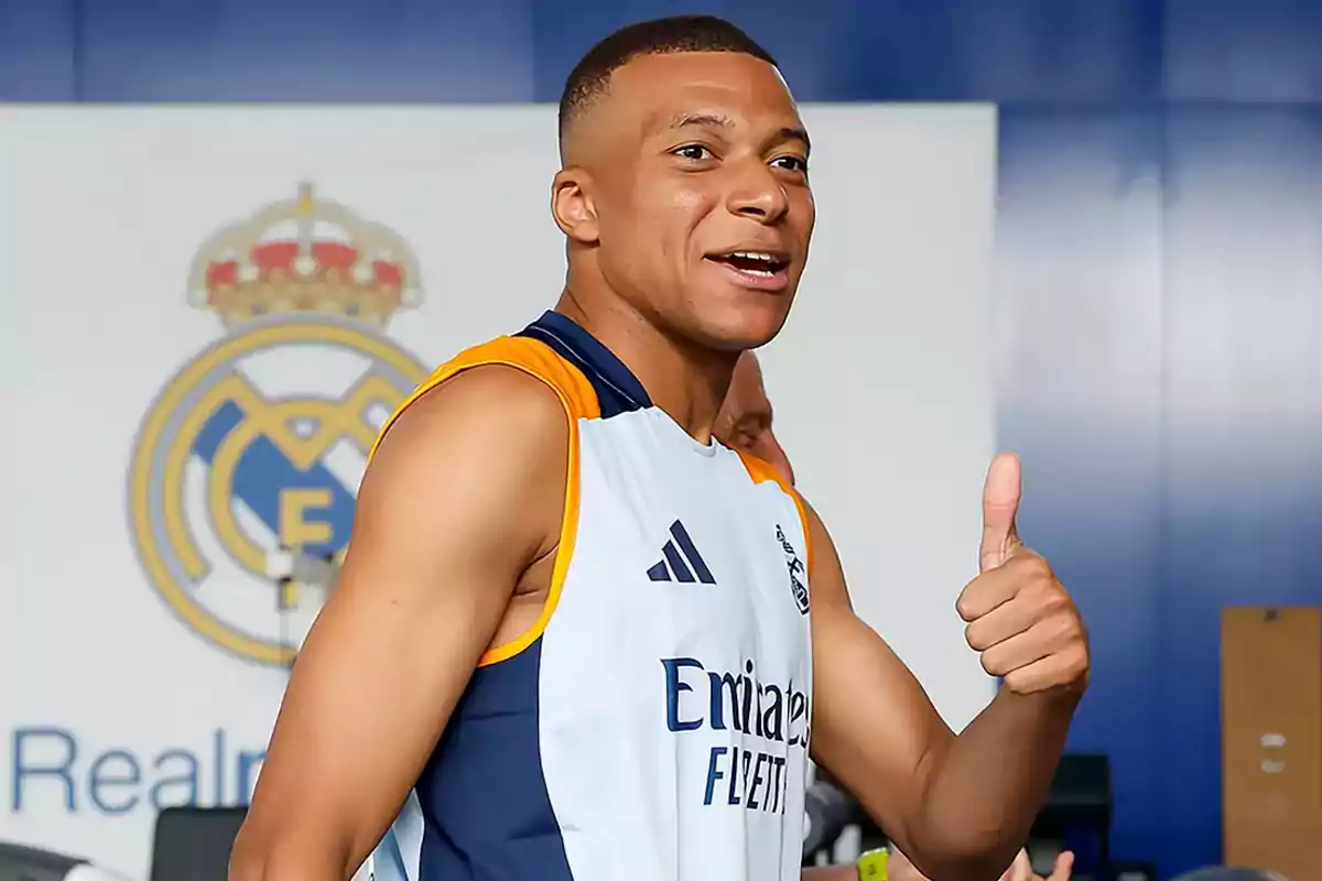 A player in a training jersey smiles and gives a thumbs up in front of a background with the Real Madrid crest.