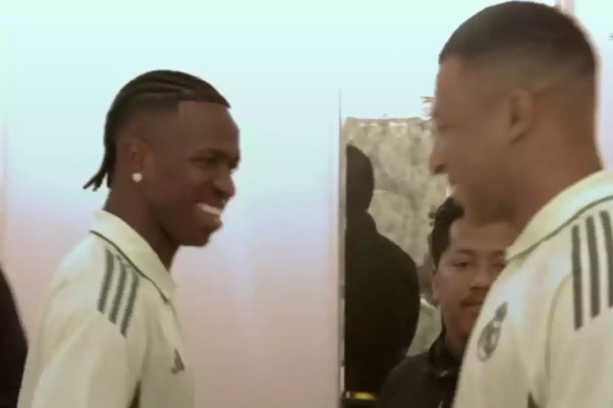 Two players in light uniforms smile at each other while meeting in a hallway, with a person watching in the background.