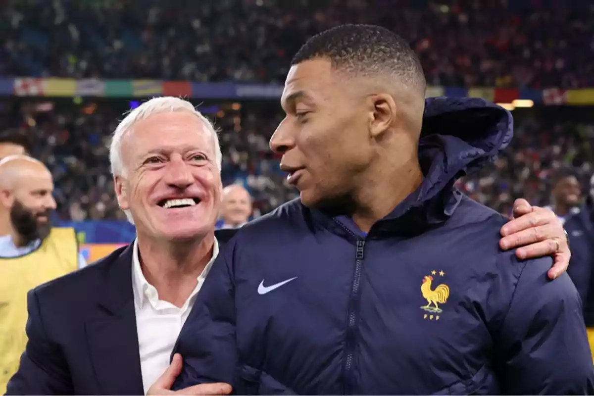 Two people smile and chat in a stadium full of spectators, one of them wearing a jacket with the French national soccer team logo. Two people smile and chat in a stadium full of spectators, one of them wearing a jacket with the French national soccer team logo.