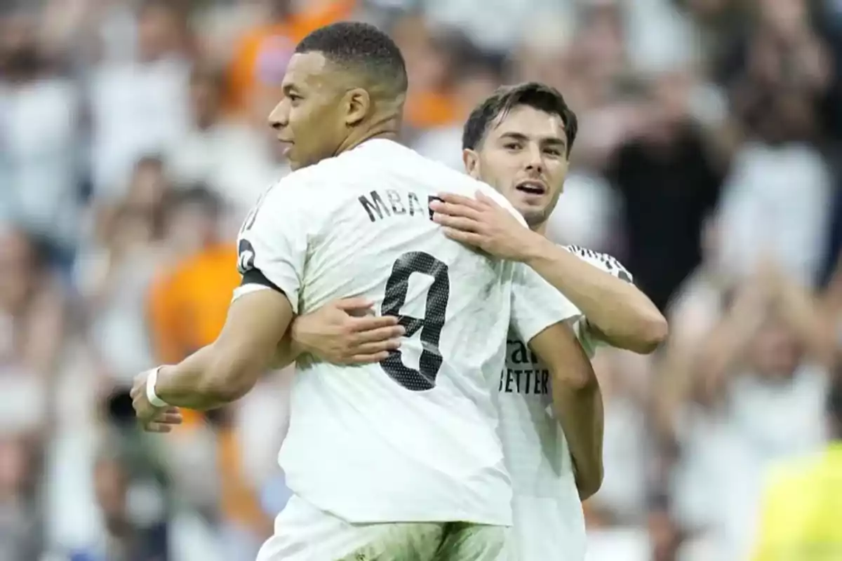 Dos jugadores de fútbol con camisetas blancas se abrazan en el campo mientras el público observa. Dos jugadores de fútbol con camisetas blancas se abrazan en el campo mientras el público observa.