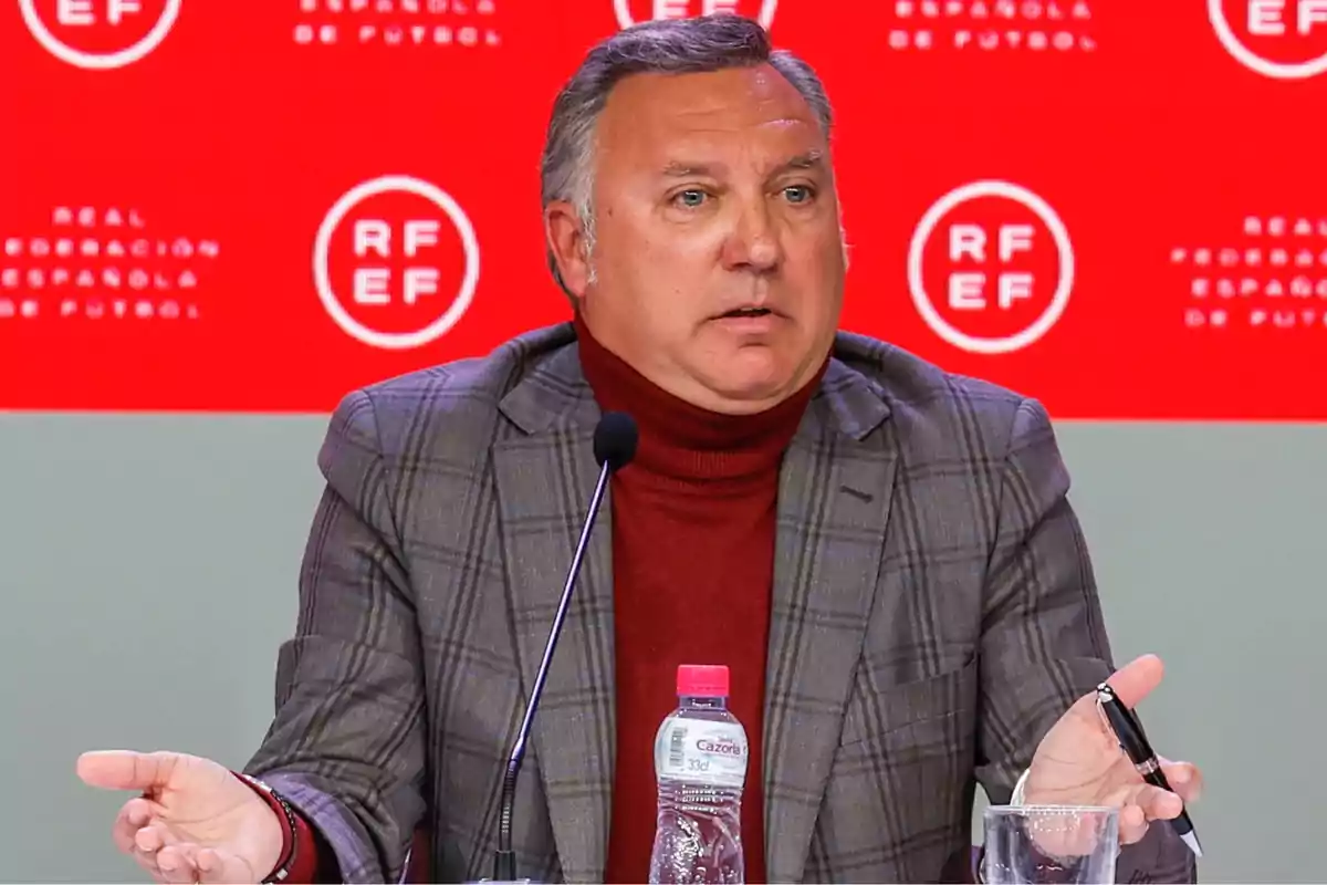 Un hombre con chaqueta a cuadros y suéter rojo habla en una conferencia de prensa frente a un fondo rojo con el logotipo de la Real Federación Española de Fútbol.