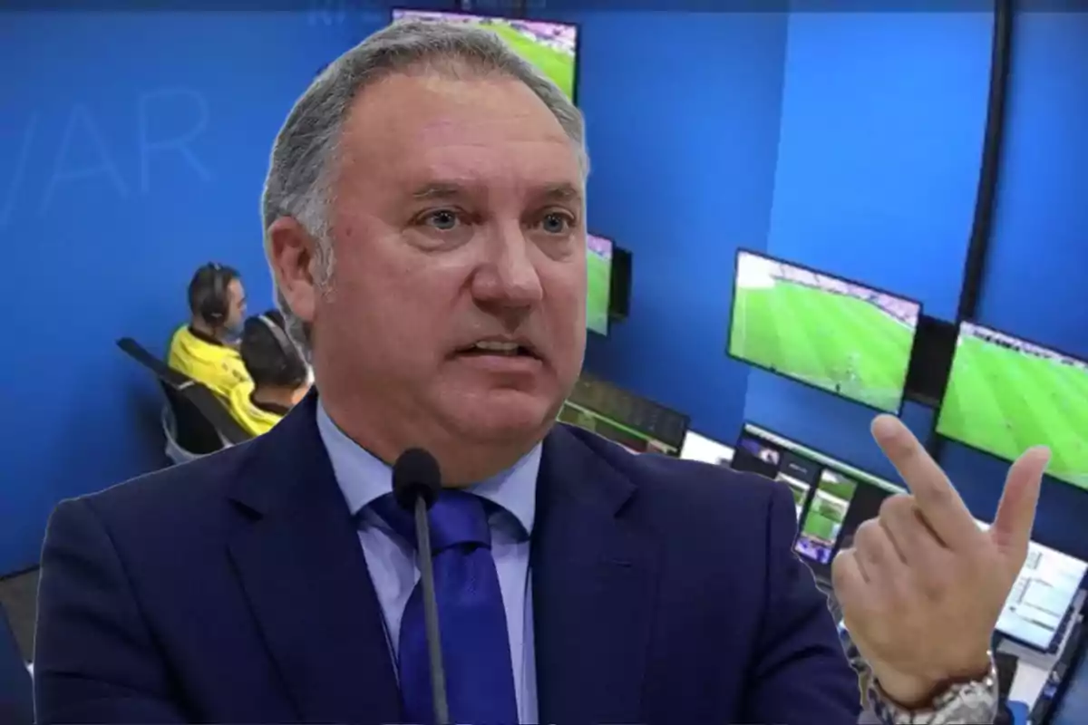 A man in a suit speaks in front of a microphone with a VAR control room in the background.