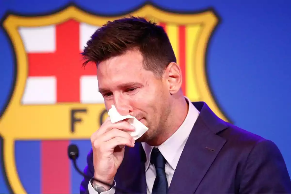 A man in a suit crying and wiping away tears with a handkerchief in front of a microphone, with a shield in the background. A man in a suit crying and wiping away tears with a handkerchief in front of a microphone, with a shield in the background.