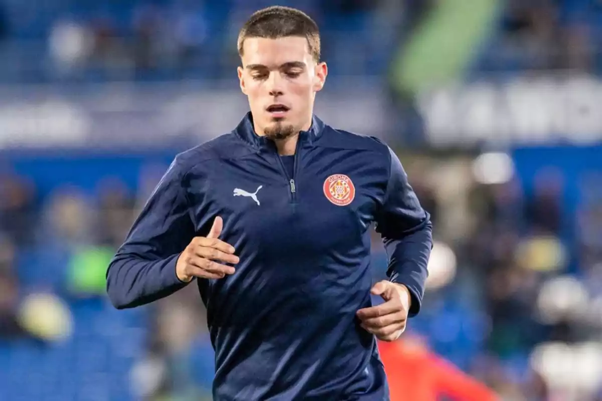 Un jugador de fútbol con uniforme azul oscuro del Girona FC corre en un estadio. Un jugador de fútbol con uniforme azul oscuro del Girona FC corre en un estadio.