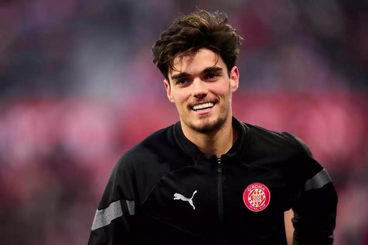 A soccer player smiles as he wears a black jacket with the Girona FC logo. A soccer player smiles as he wears a black jacket with the Girona FC logo.