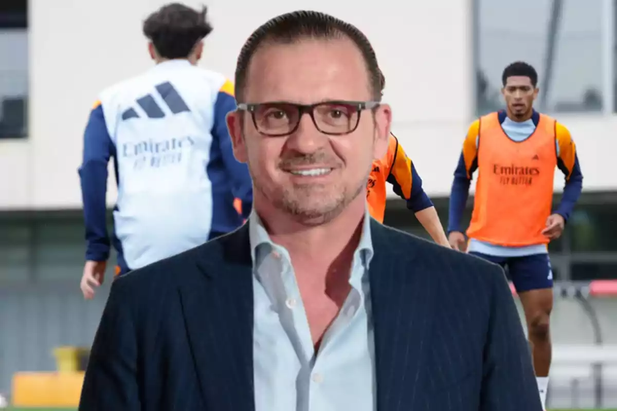 A man in glasses and a suit smiles in front of a soccer training field where players in orange bibs can be seen.