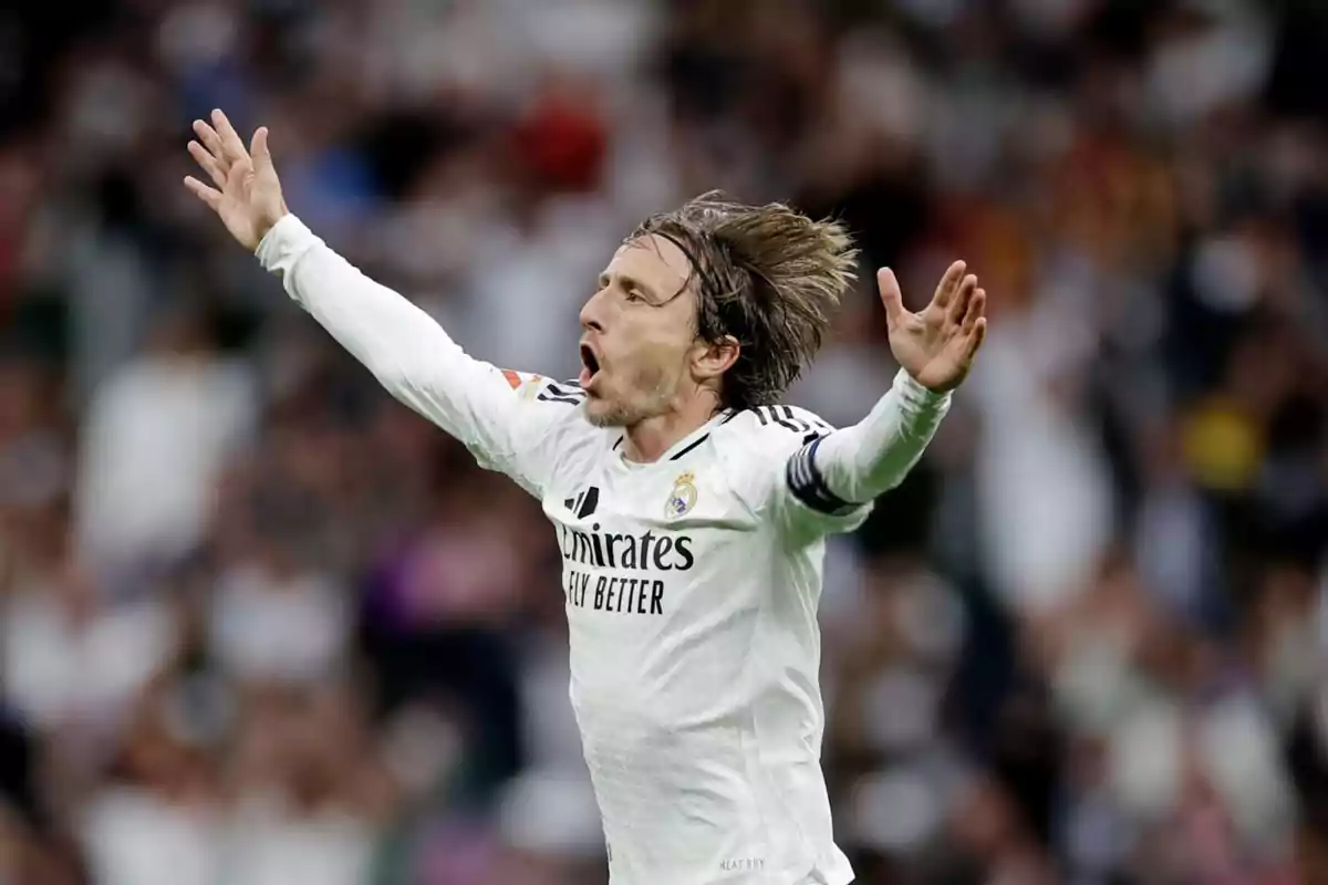 Un jugador de fútbol con uniforme blanco celebra con los brazos abiertos en un estadio.