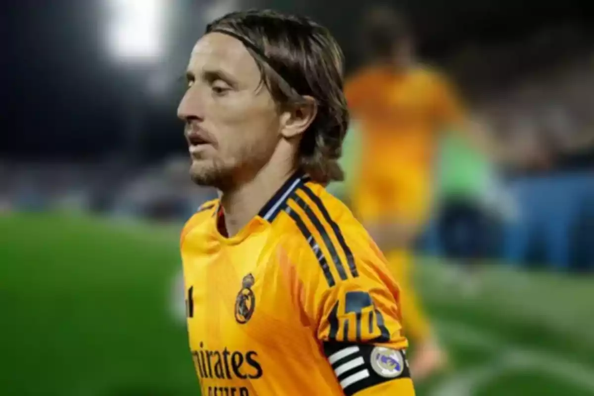 Un futbolista con uniforme naranja y brazalete de capitán en un campo de fútbol.