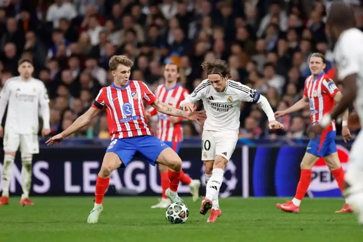 Two players compete for the ball in a match, one wearing a white uniform and the other a red and white striped uniform, while other players and spectators watch in the background.