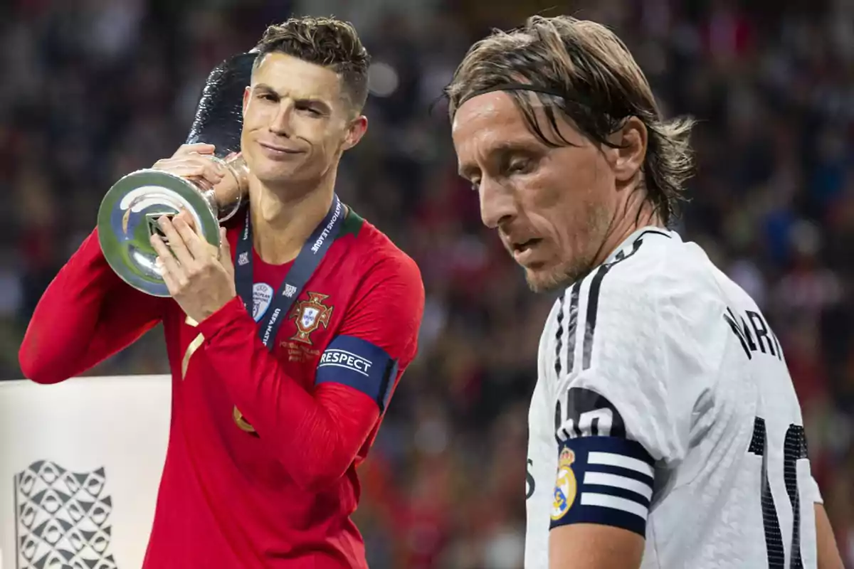 Dos futbolistas en el campo, uno con uniforme rojo sosteniendo un trofeo y el otro con uniforme blanco.