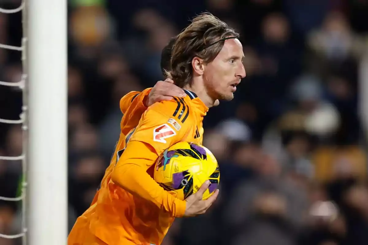 A soccer player in an orange uniform stands on the playing field and holds the ball.