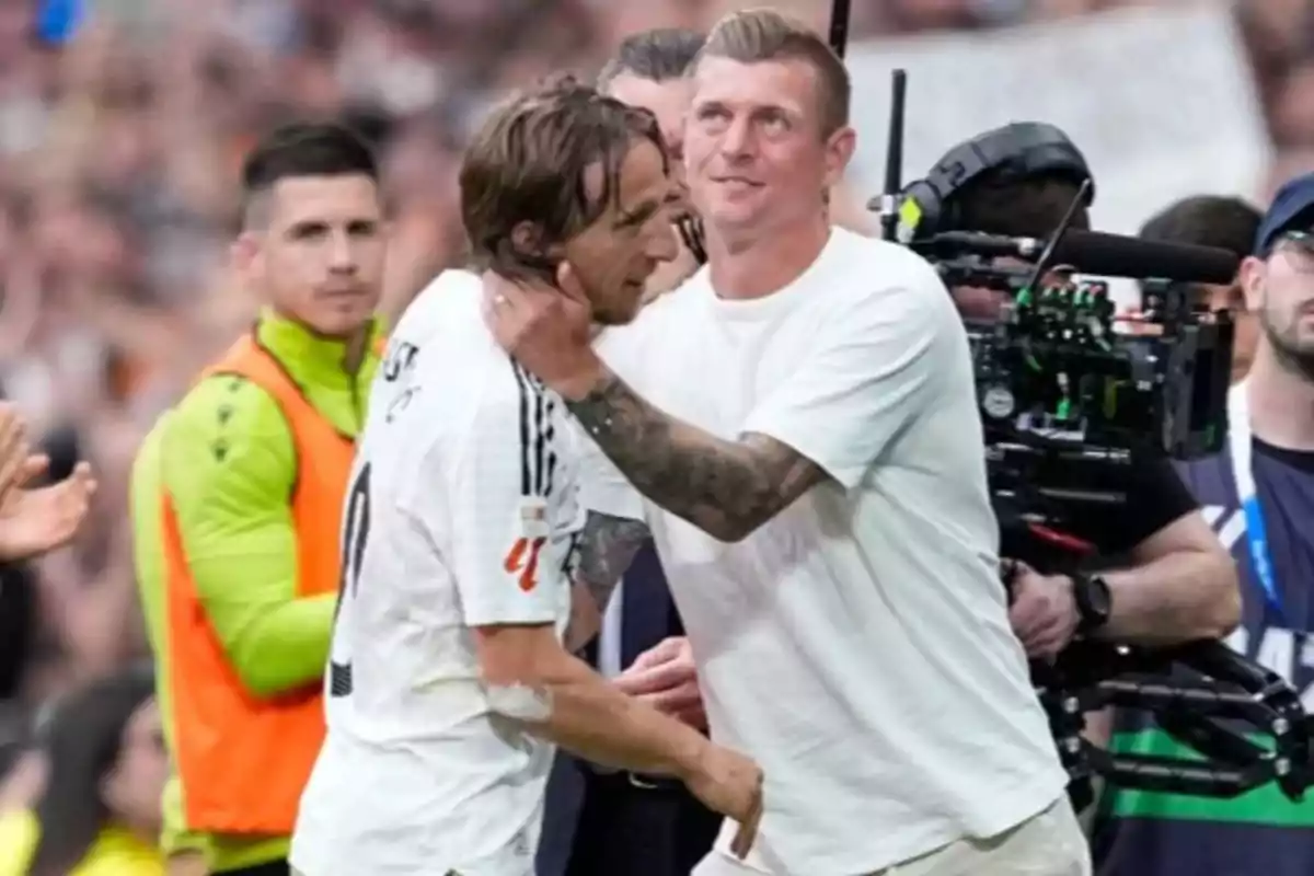 A player hugs another on the field while a camera records the moment and several people watch in the background. A player hugs another on the field while a camera records the moment and several people watch in the background.