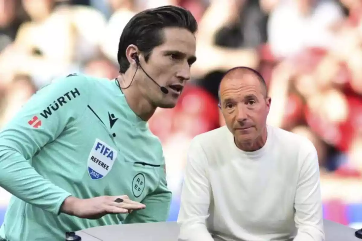 Un árbitro de fútbol con uniforme verde y auricular junto a un hombre calvo con suéter blanco.