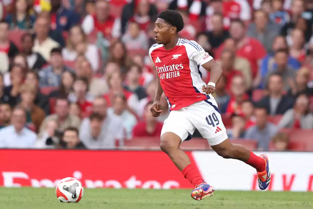 Arsenal player running on the field during a match with the ball nearby and fans in the stands in the background Arsenal player running on the field during a match with the ball nearby and fans in the stands in the background