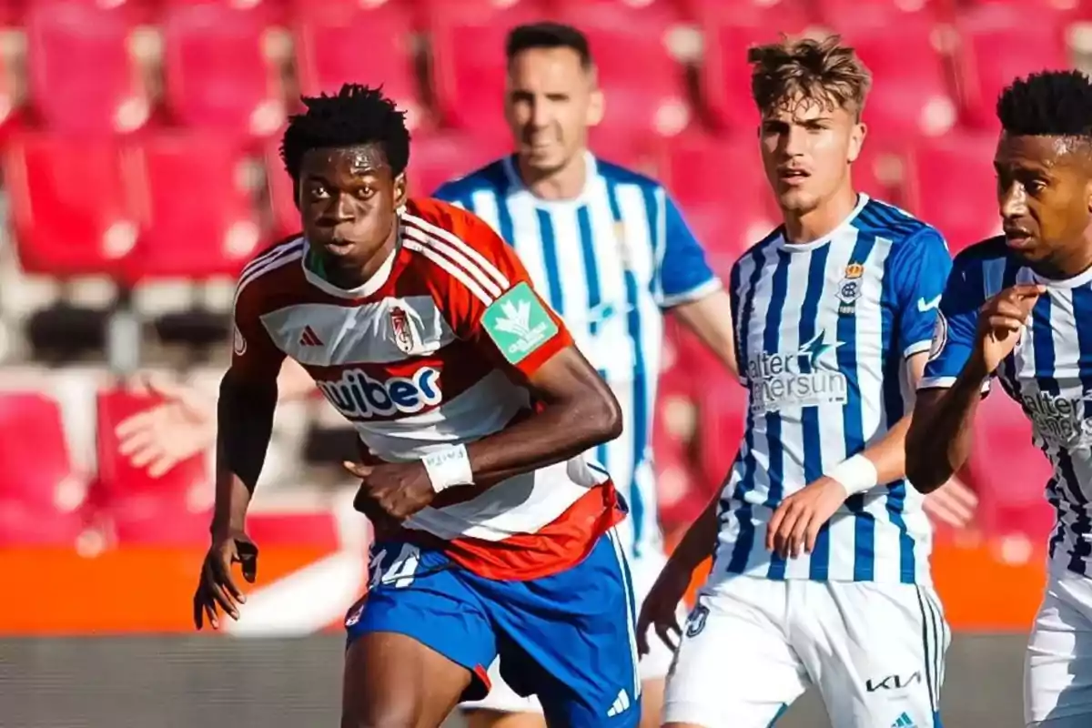 Players from two soccer teams compete for the ball in a match with red stands in the background. Players from two soccer teams compete for the ball in a match with red stands in the background.
