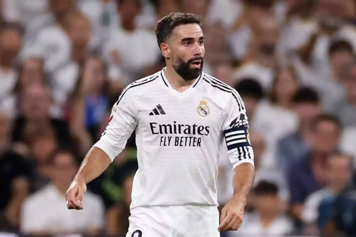 Un jugador de fútbol con uniforme blanco y brazalete de capitán en un estadio lleno de espectadores.