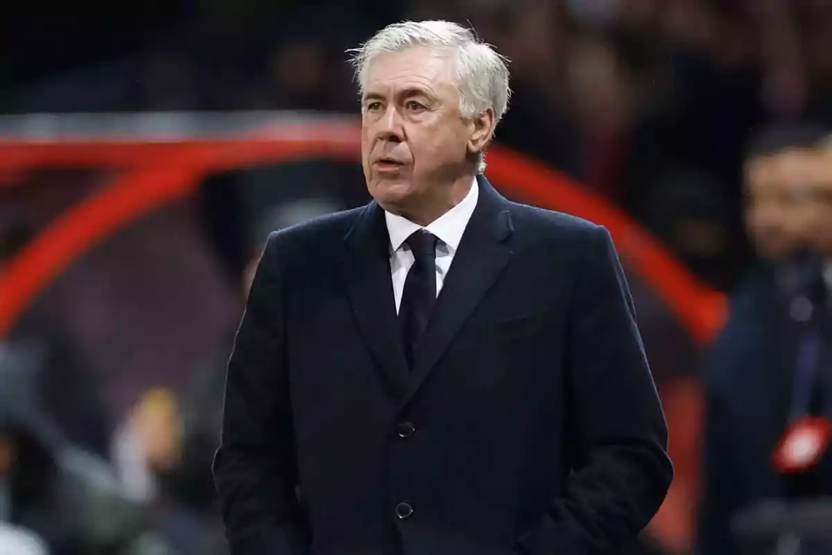 A gray-haired man in a dark suit is standing in a sports setting.