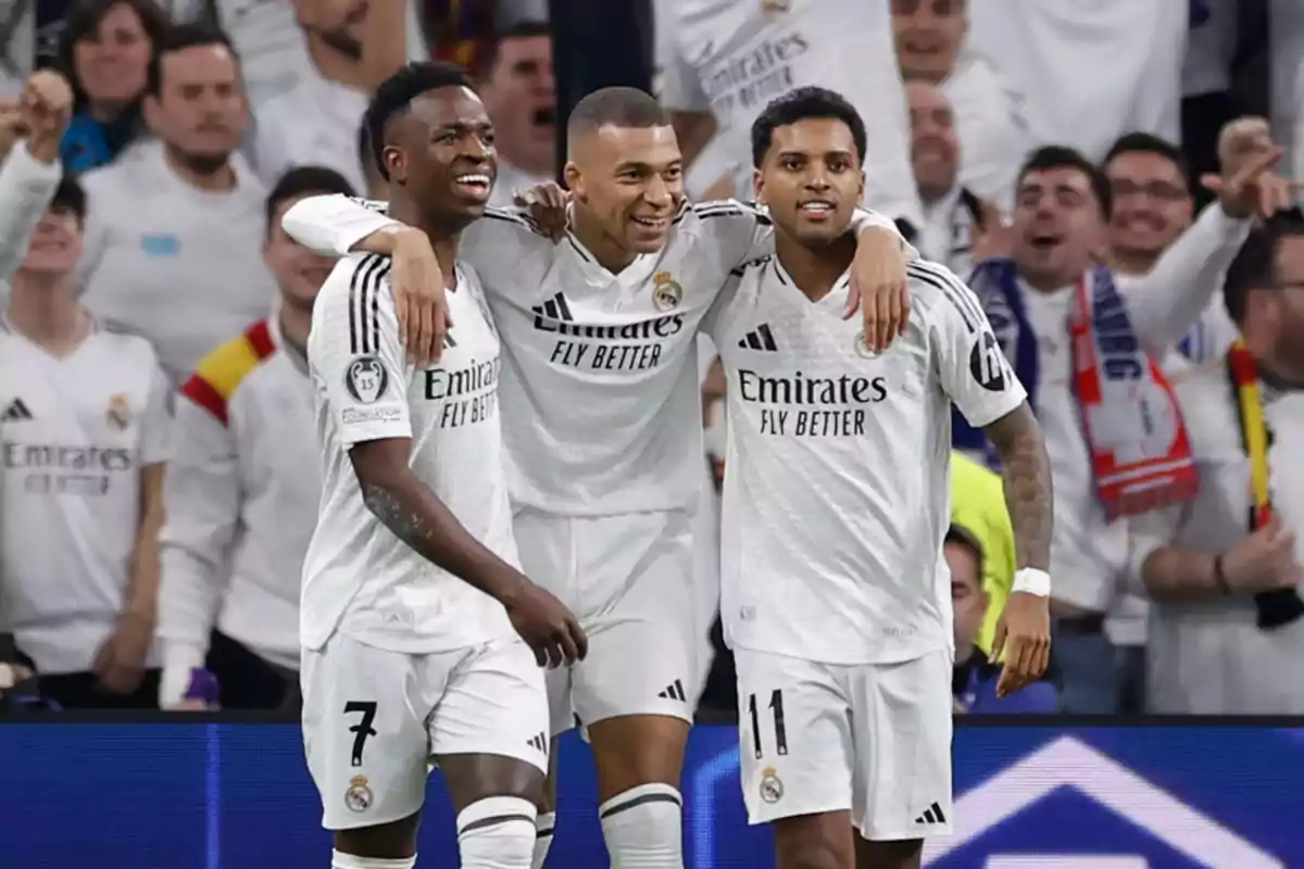 Tres jugadores de fútbol con uniformes blancos celebran en el campo mientras el público los anima.