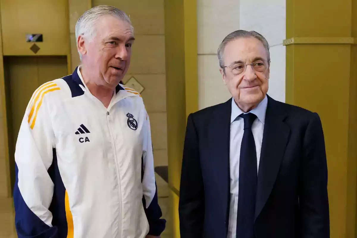 Dos hombres conversando en un edificio, uno lleva un chándal blanco con detalles en azul y amarillo y el otro viste un traje oscuro.