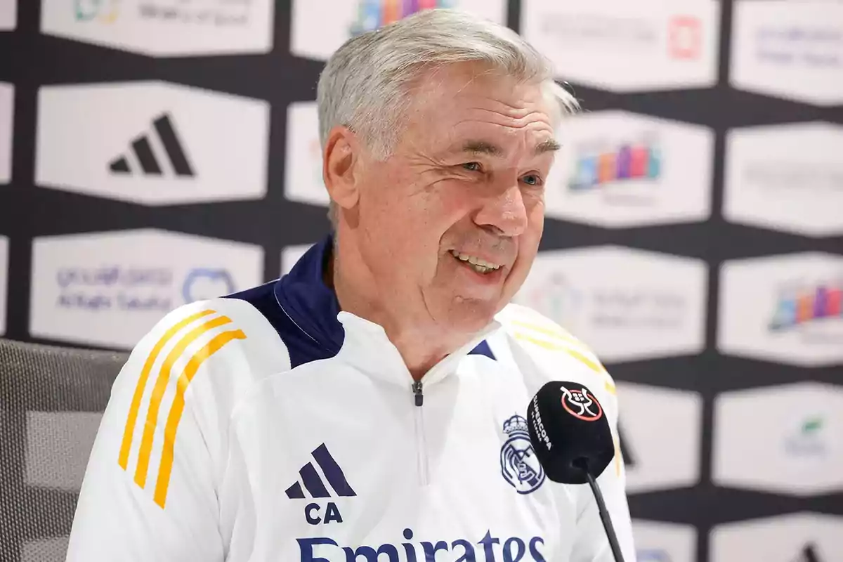A man with grey hair and a white Real Madrid jacket smiles as he speaks at a news conference. A man with grey hair and a white Real Madrid jacket smiles as he speaks at a news conference.