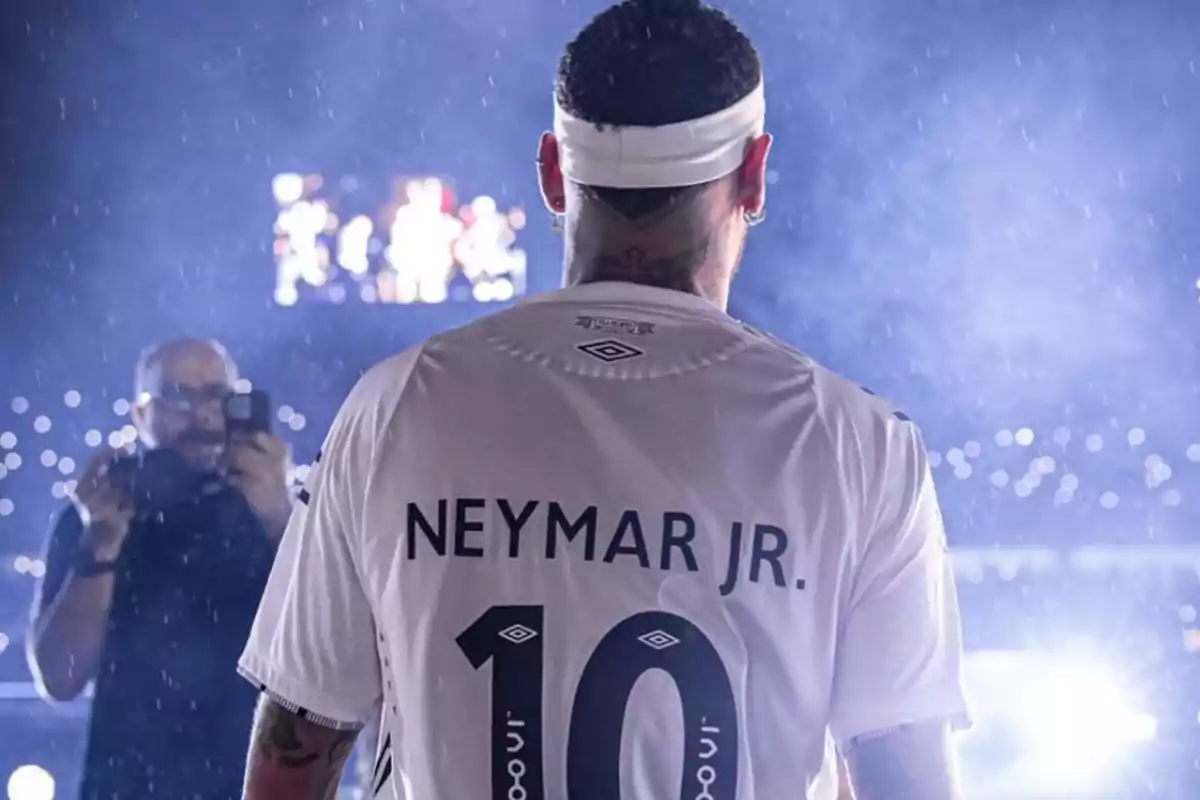 Un jugador de fútbol de espaldas con una camiseta blanca y el número 10, mientras un fotógrafo lo captura bajo la lluvia en un estadio iluminado. Un jugador de fútbol de espaldas con una camiseta blanca y el número 10, mientras un fotógrafo lo captura bajo la lluvia en un estadio iluminado.
