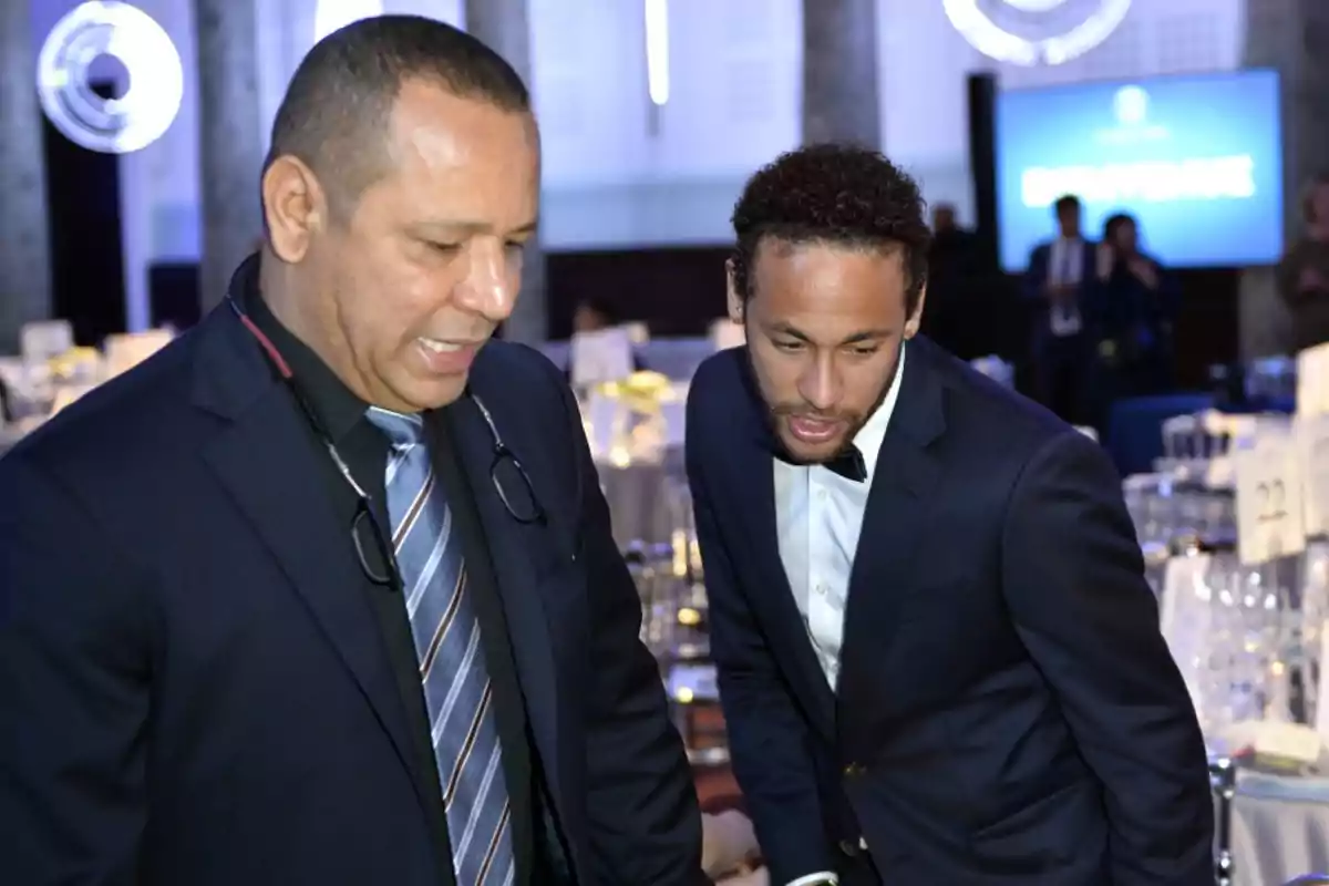 Two men dressed in suits are talking at a formal event with tables and glasses in the background.