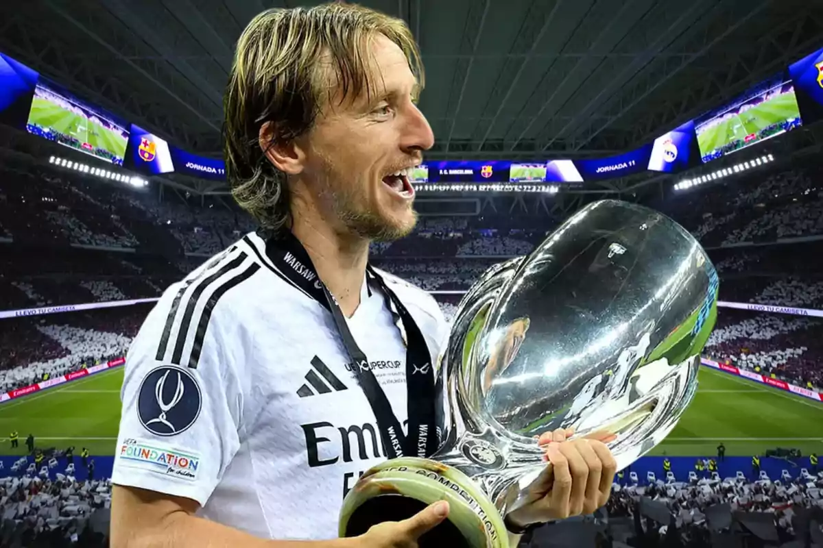 Un jugador de fútbol con uniforme blanco sostiene un trofeo grande y plateado en un estadio lleno de espectadores.