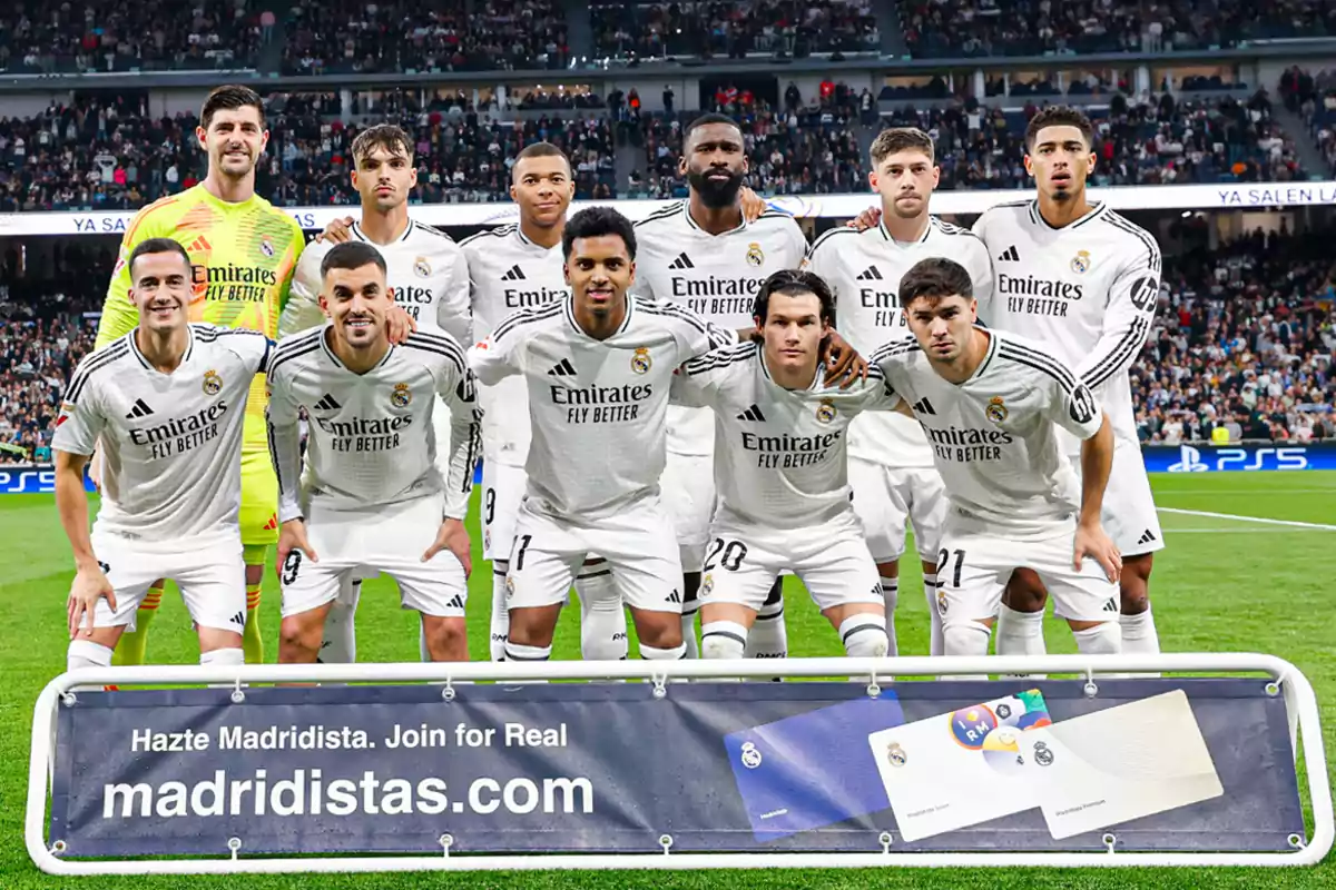 A soccer team posing on the field in white jerseys with a promotional poster in front.