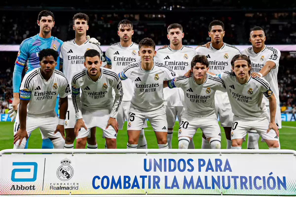 A soccer team poses on the field in white jerseys, behind a banner promoting the fight against malnutrition.