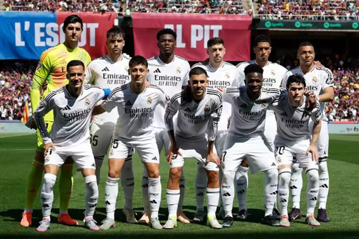 Un equipo de fútbol posando en el campo antes de un partido, con jugadores vistiendo uniformes blancos y un estadio lleno de espectadores al fondo.