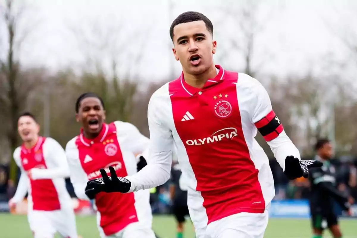 Jugador del Ajax celebrando un gol en el campo con compañeros al fondo mostrando emoción Jugador del Ajax celebrando un gol en el campo con compañeros al fondo mostrando emoción