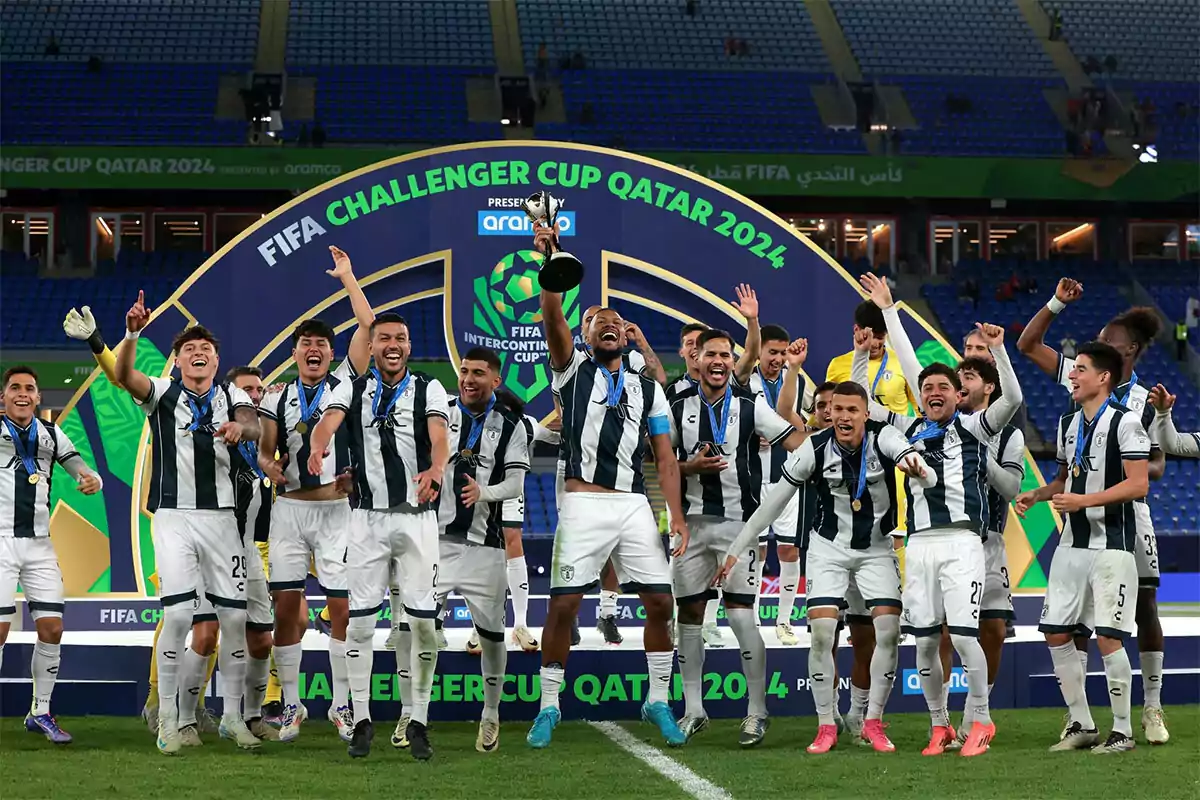 Un equipo de fútbol celebra con entusiasmo en el campo mientras uno de los jugadores levanta un trofeo, todos visten uniformes a rayas blancas y negras y llevan medallas alrededor del cuello, al fondo se ve un cartel que dice FIFA Challenger Cup Qatar 2024.