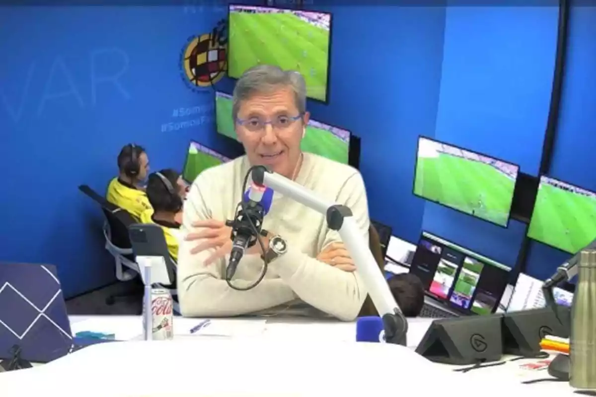 Un hombre en un estudio de radio con varios monitores mostrando un partido de fútbol en el fondo.