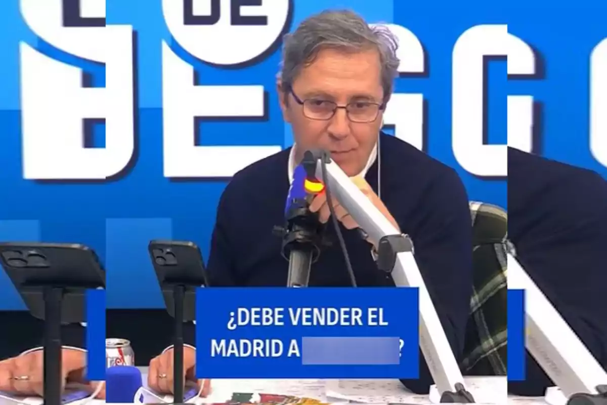 A man with glasses and a dark sweater is sitting in front of a microphone in a radio studio, with a blue sign that has a question about Real Madrid.