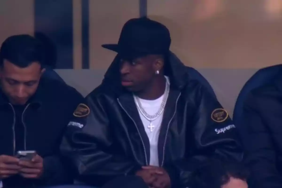 Two people sitting, one looking at his phone and the other wearing a black jacket and cap, looking to the side. Two people sitting, one looking at his phone and the other wearing a black jacket and cap, looking to the side.