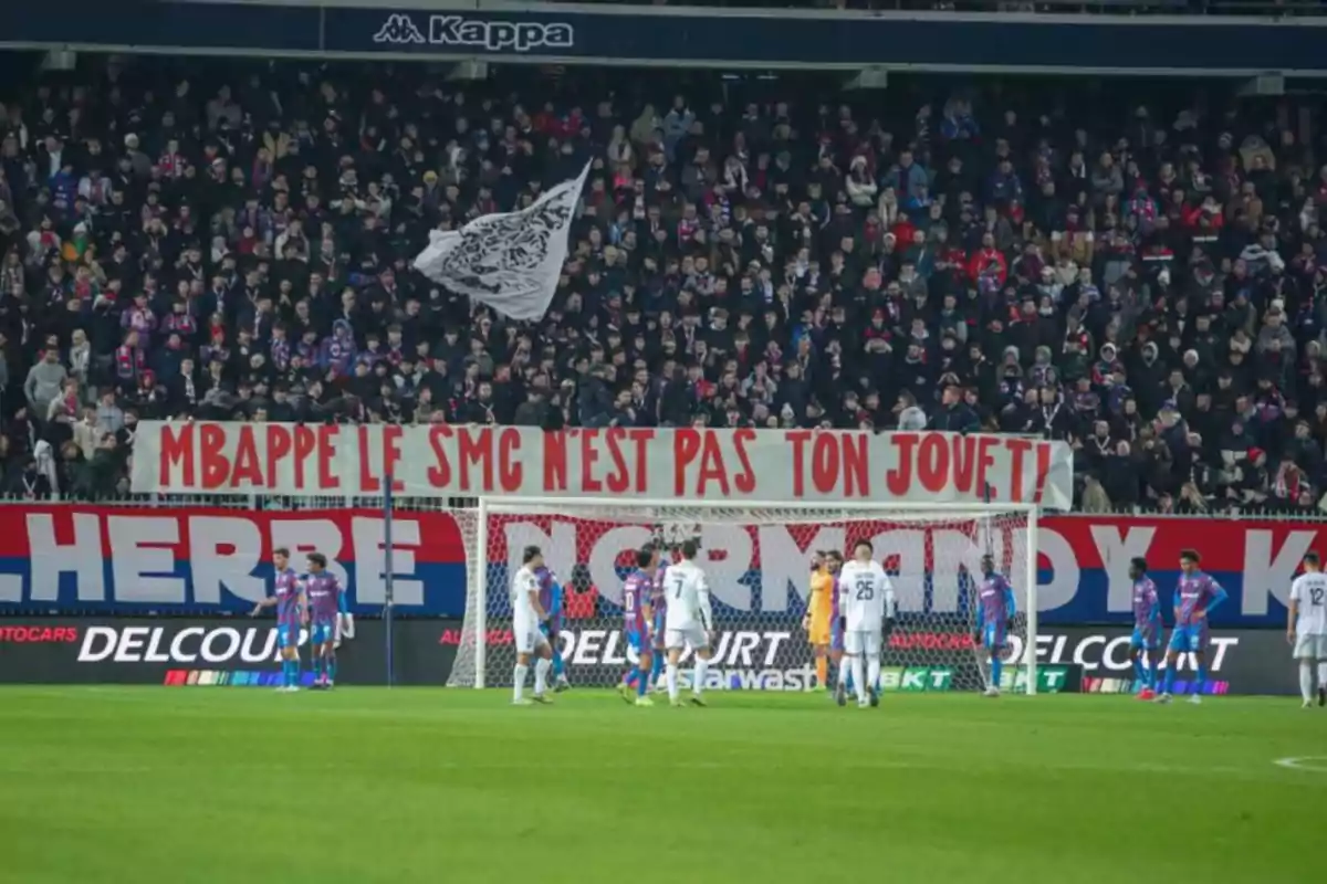 Un grupo de aficionados en un estadio de fútbol sostiene una pancarta con un mensaje dirigido a un jugador mientras los equipos están en el campo. Un grupo de aficionados en un estadio de fútbol sostiene una pancarta con un mensaje dirigido a un jugador mientras los equipos están en el campo.