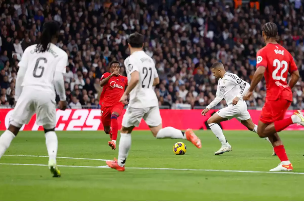 Players from two different soccer teams compete for the ball in a match, with a stadium full of spectators in the background. Players from two different soccer teams compete for the ball in a match, with a stadium full of spectators in the background.