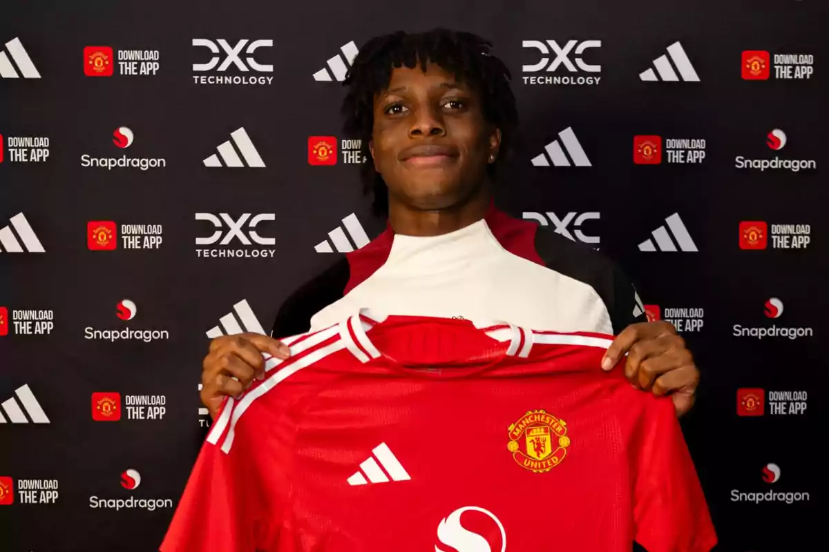 Una persona sosteniendo una camiseta roja del Manchester United frente a un fondo con logotipos de DXC Technology, Snapdragon y Adidas. Una persona sosteniendo una camiseta roja del Manchester United frente a un fondo con logotipos de DXC Technology, Snapdragon y Adidas.