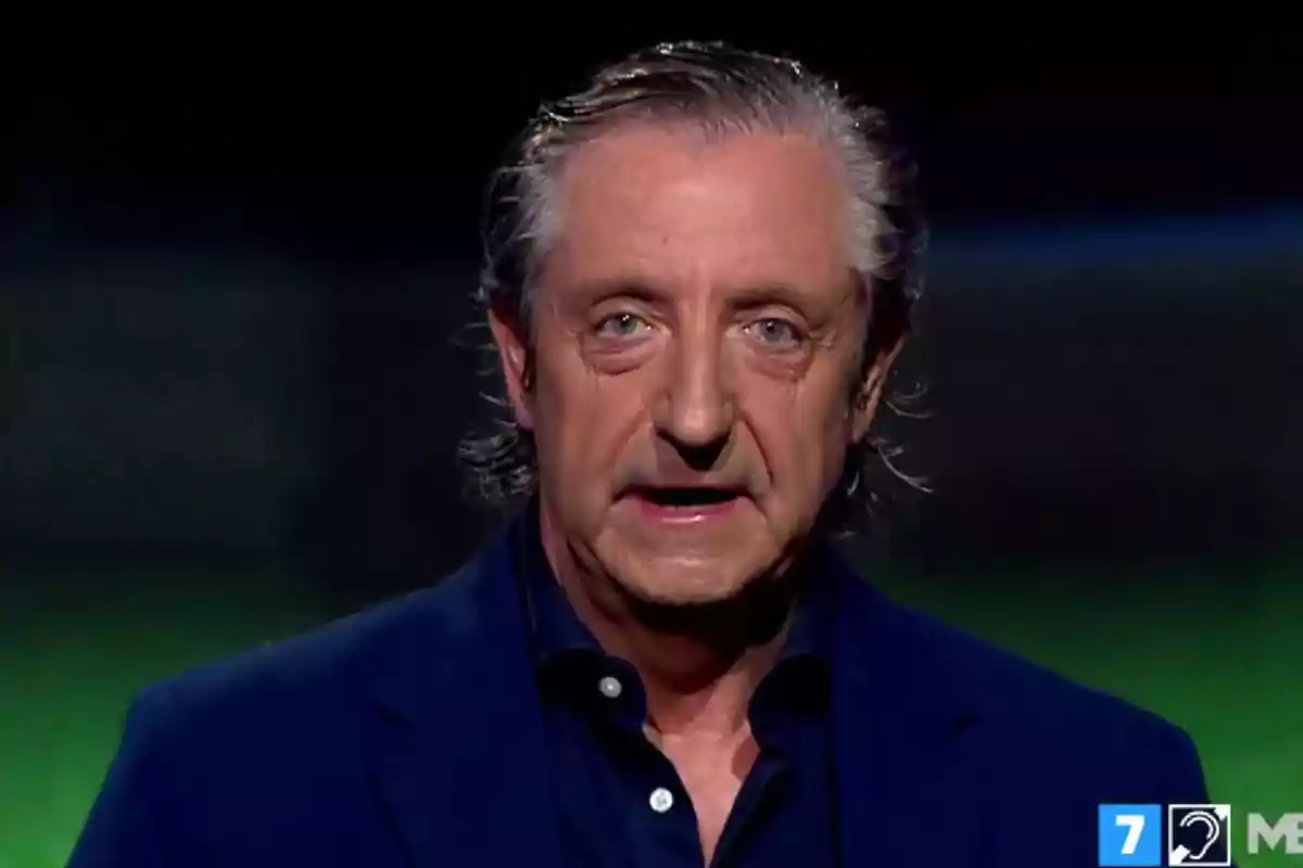 Gray-haired man with a serious expression wearing a blue jacket in a television studio