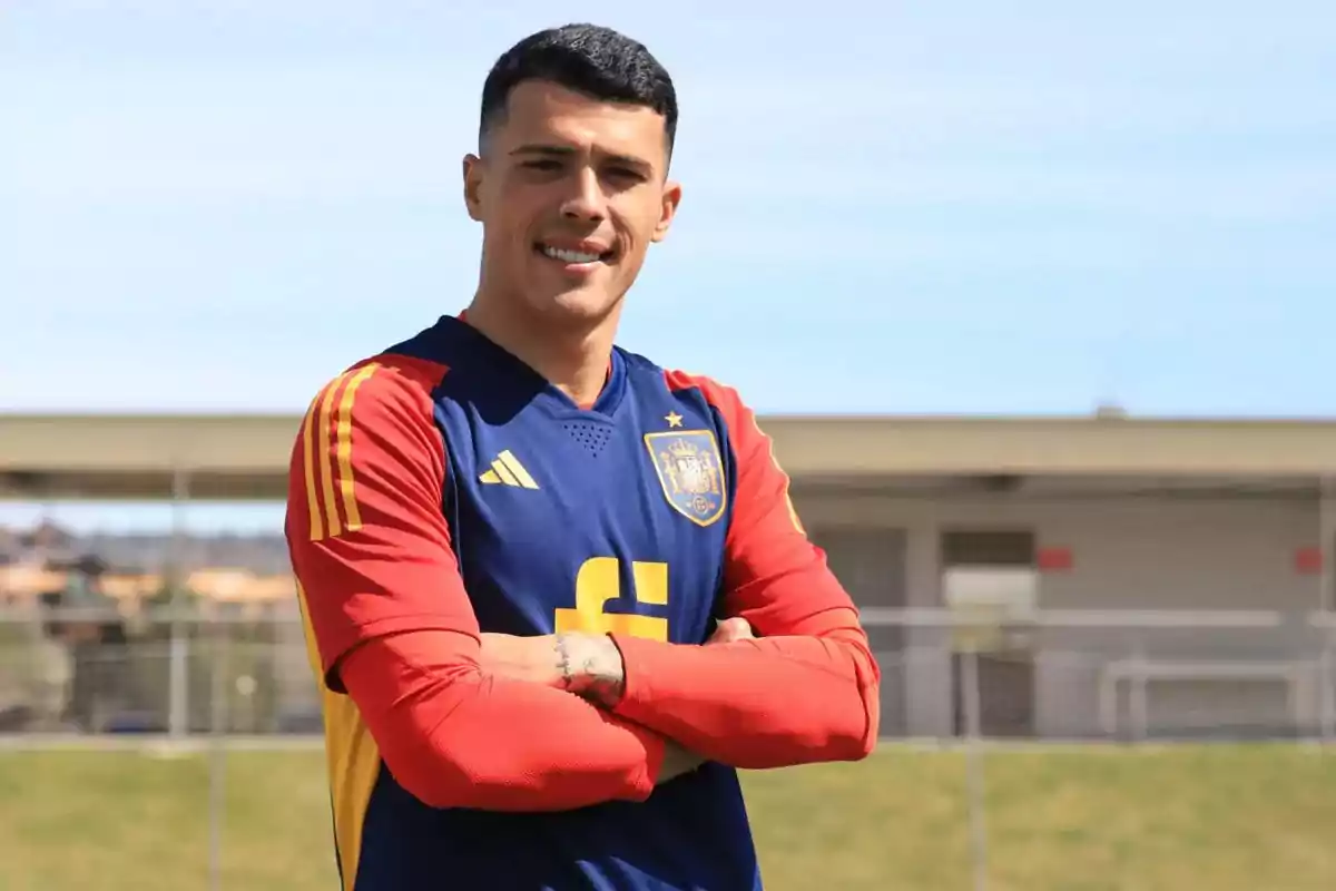 Un hombre con uniforme de fútbol de España sonríe con los brazos cruzados en un campo deportivo. Un hombre con uniforme de fútbol de España sonríe con los brazos cruzados en un campo deportivo.