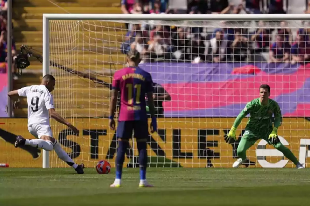 Soccer player in a white uniform kicking a ball toward the goal while the goalkeeper and another player watch in a stadium full of spectators.