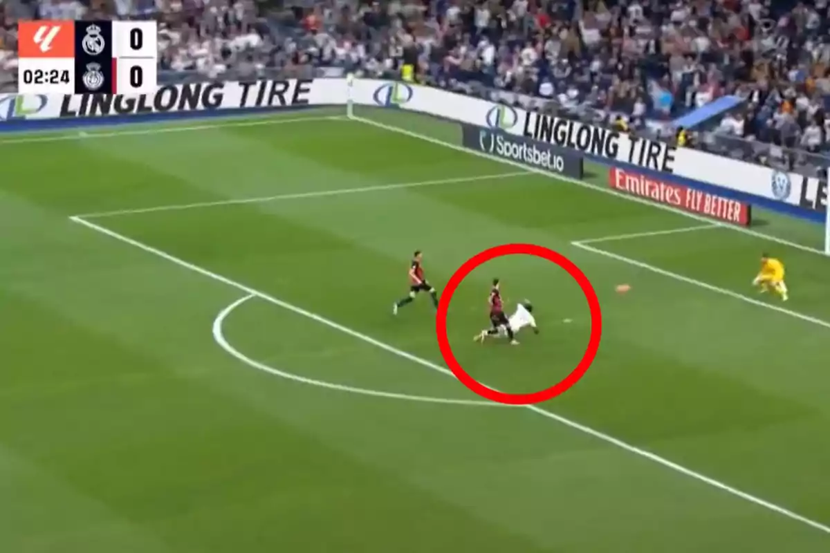 Un jugador de fútbol cae al suelo dentro del área mientras otro lo sigue de cerca, con el portero atento a la jugada y el marcador mostrando un empate a cero.