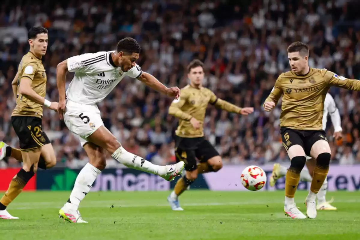 A player in a white uniform kicks the ball while other players in gold uniforms watch him in a stadium full of spectators. A player in a white uniform kicks the ball while other players in gold uniforms watch him in a stadium full of spectators.