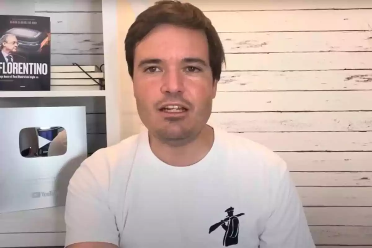 Un hombre con camiseta blanca está frente a una pared de madera clara, a su lado hay una estantería con un libro y una placa de YouTube.