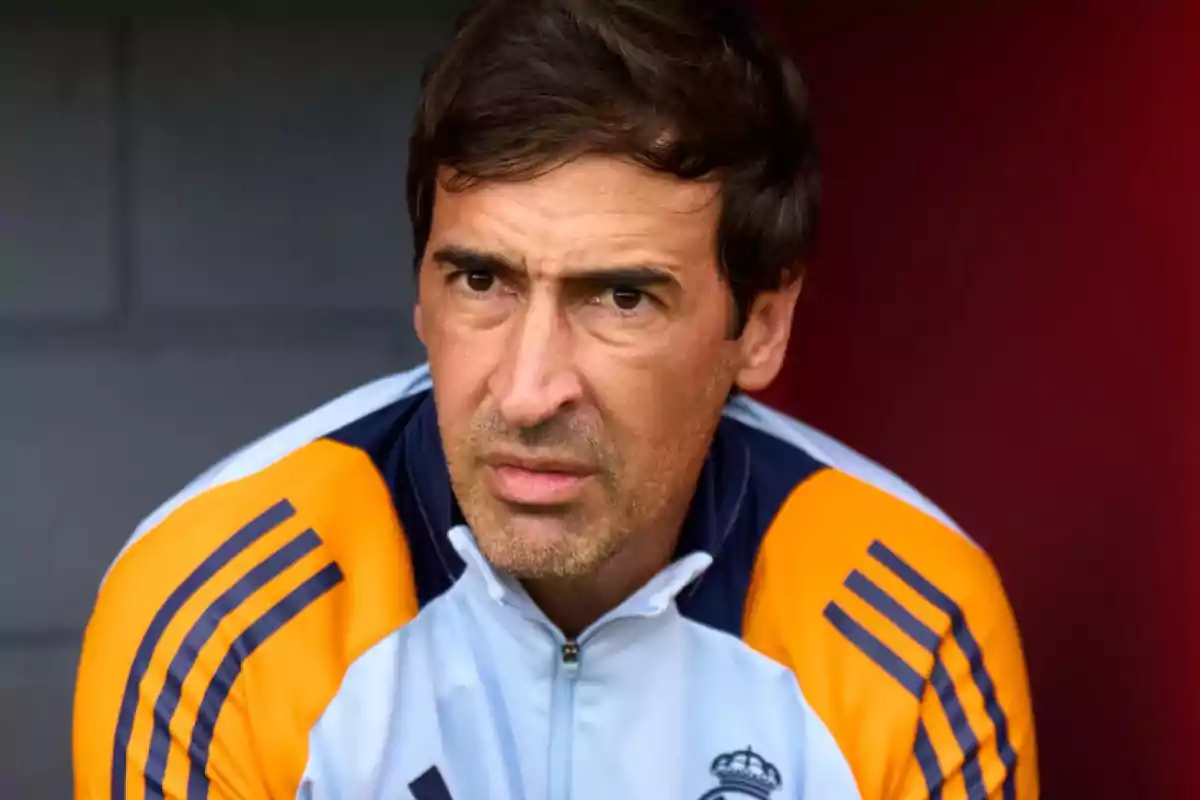 Hombre con chaqueta deportiva blanca y naranja con el escudo del Real Madrid mirando al frente con expresión seria Hombre con chaqueta deportiva blanca y naranja con el escudo del Real Madrid mirando al frente con expresión seria