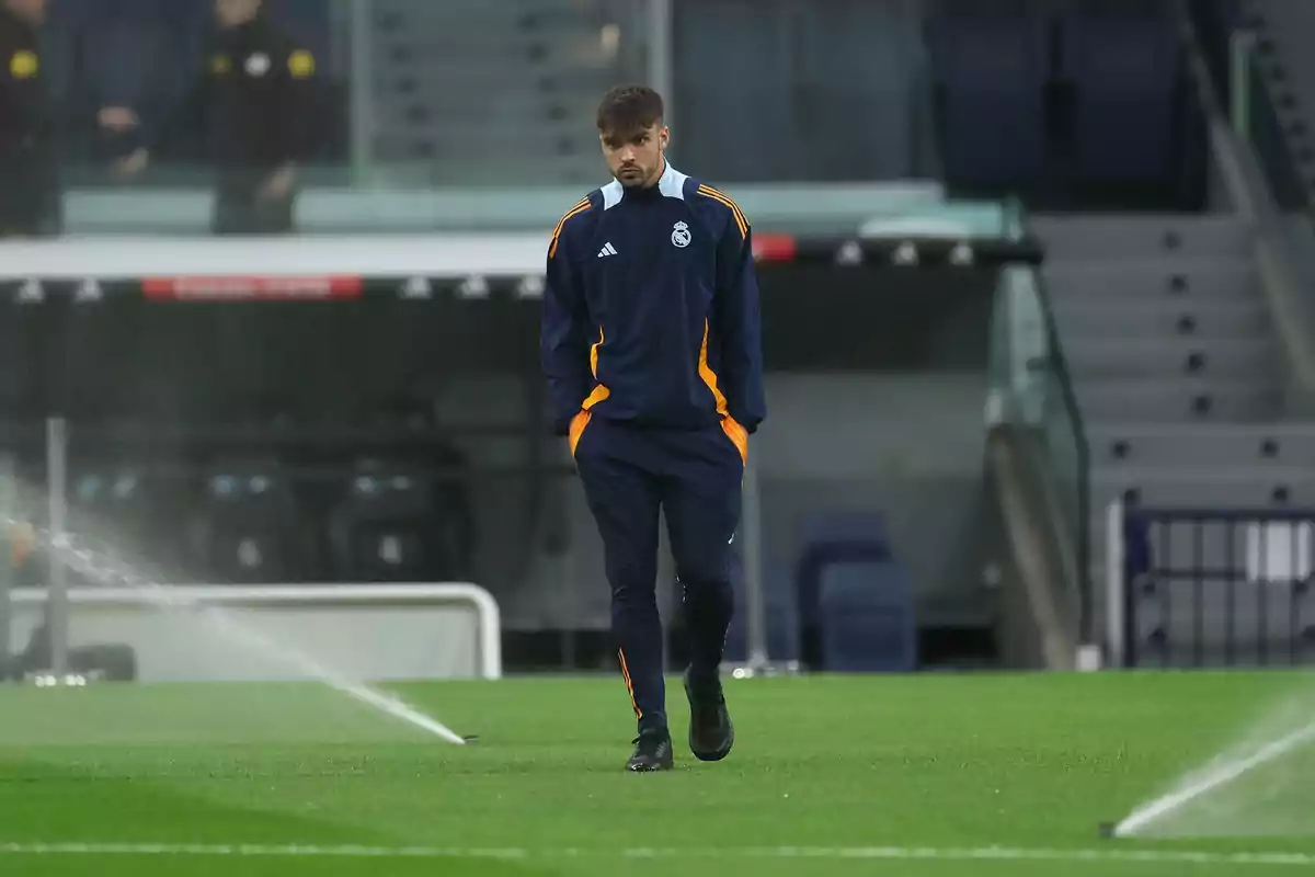 Jugador con chándal azul marino del Real Madrid caminando por el campo de fútbol mientras los aspersores riegan el césped Jugador con chándal azul marino del Real Madrid caminando por el campo de fútbol mientras los aspersores riegan el césped