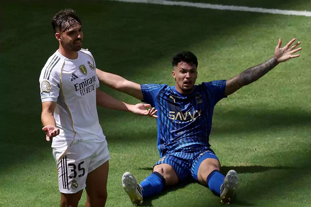 Un jugador de fútbol con uniforme blanco de pie y otro con uniforme azul sentado en el césped levantando los brazos, ambos mostrando gestos de inconformidad durante un partido. Un jugador de fútbol con uniforme blanco de pie y otro con uniforme azul sentado en el césped levantando los brazos, ambos mostrando gestos de inconformidad durante un partido.