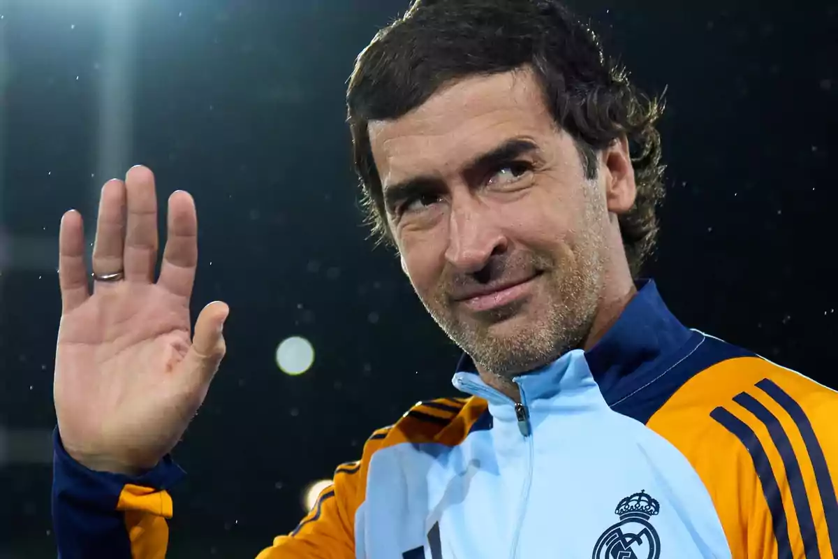 Hombre con chaqueta deportiva del Real Madrid saludando con la mano y sonriendo frente a un fondo oscuro