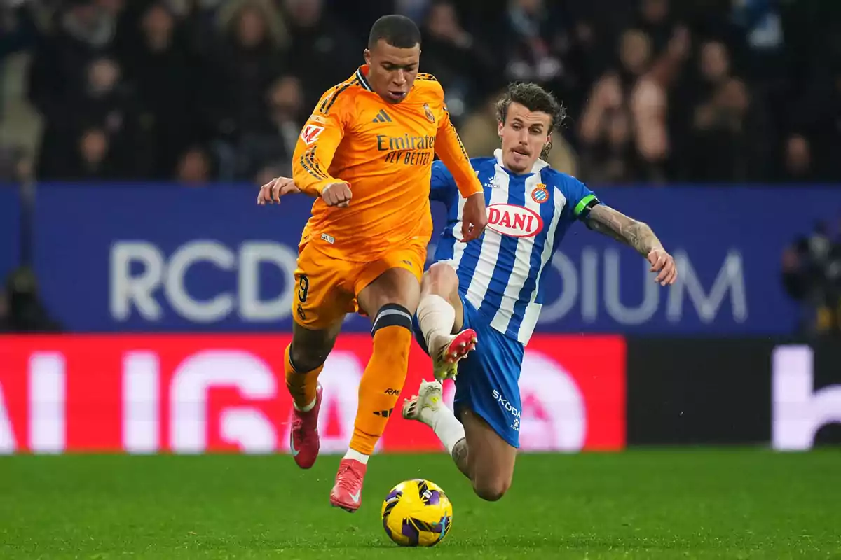 Dos jugadores de fútbol compiten por el balón en un partido, uno viste de naranja y el otro de azul y blanco, en un estadio con público de fondo.