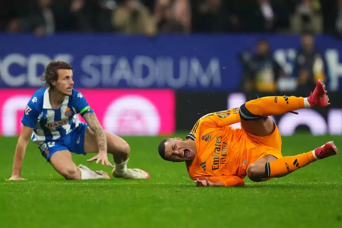 Un jugador de fútbol con uniforme naranja está en el suelo con expresión de dolor mientras otro jugador con uniforme azul y blanco lo observa desde cerca en un campo de fútbol.